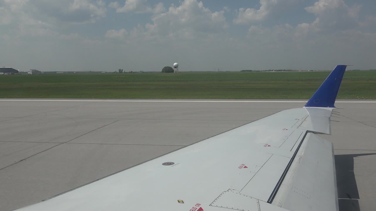 Passenger-window view in Winnipeg, Canada, showing a white airplane wing with a blue winglet over gray runway pavement, green airfield grass, distant structures, clouds, and no visible people.