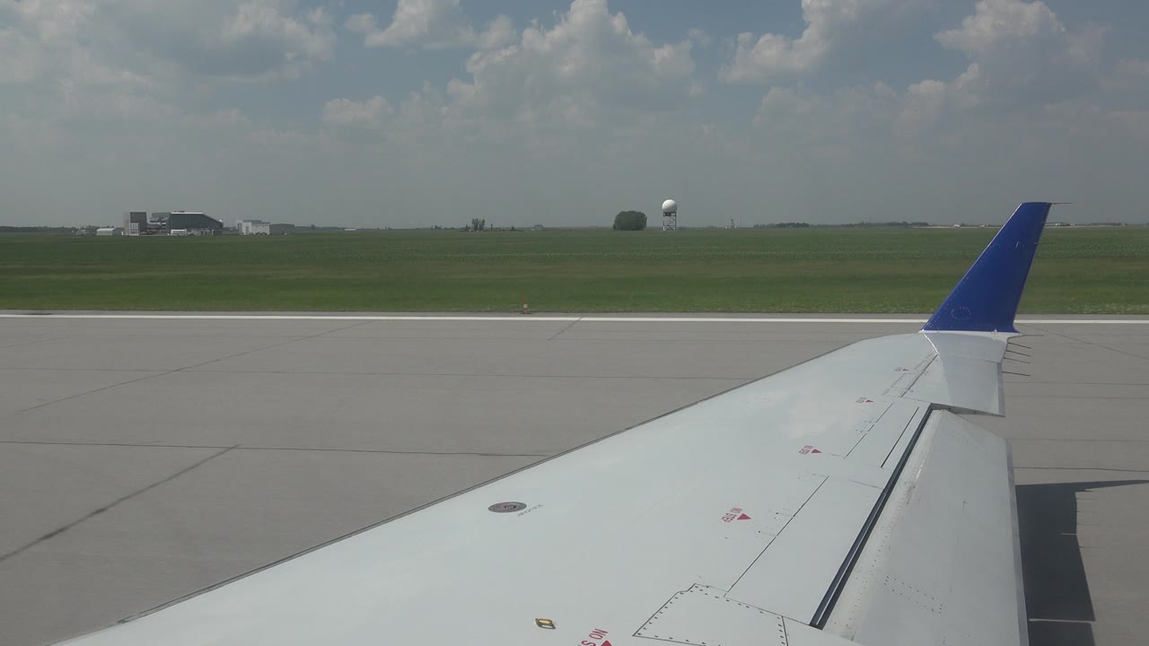 Passenger-window view of a white airplane wing and blue winglet over runway pavement in Winnipeg, Canada, with grassy airfield, distant structures, clouds, and no visible people.
