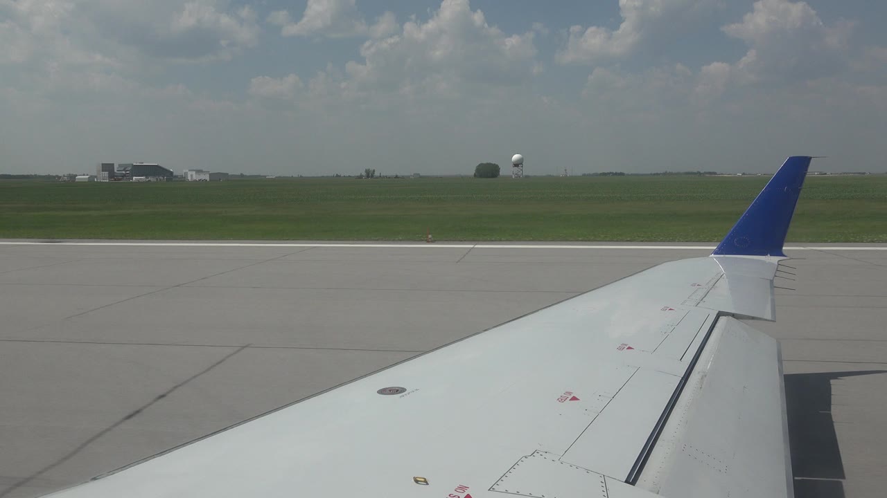 White aircraft wing angles across the foreground as the runway and green airfield pass behind it in Winnipeg, Canada, with low buildings on the horizon and no visible people.