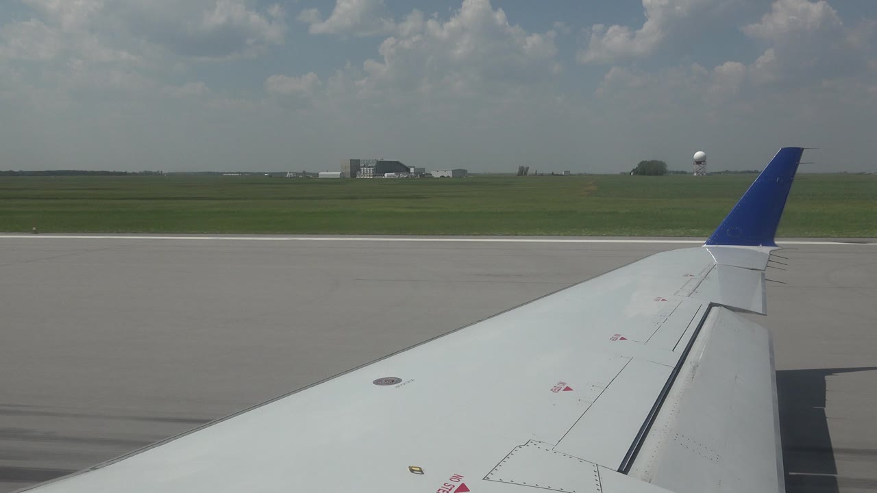 White airplane wing with a blue tip seen from a passenger-window angle on a Winnipeg runway, with green fields, distant airport buildings, clouds, and no visible people.