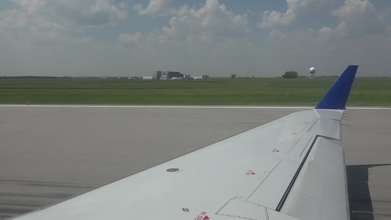 Passenger-window view of the white wing and blue wingtip on the Winnipeg runway, with more pavement visible beside the wing, low airport structures, and no visible people.