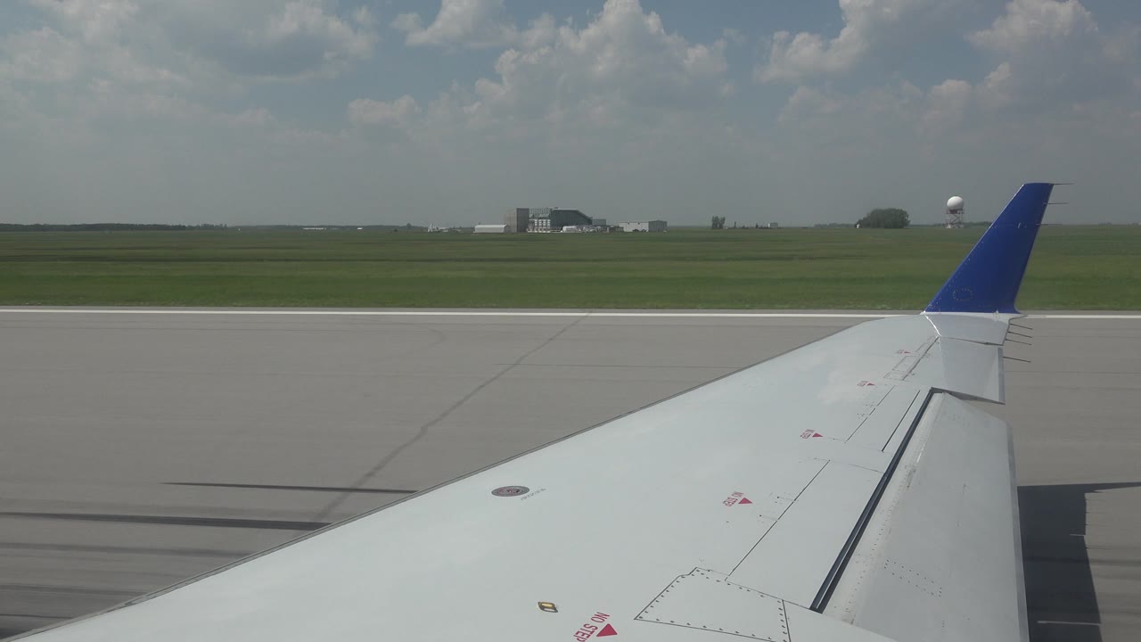 Blue-tipped airplane wing passing runway markings and open field.