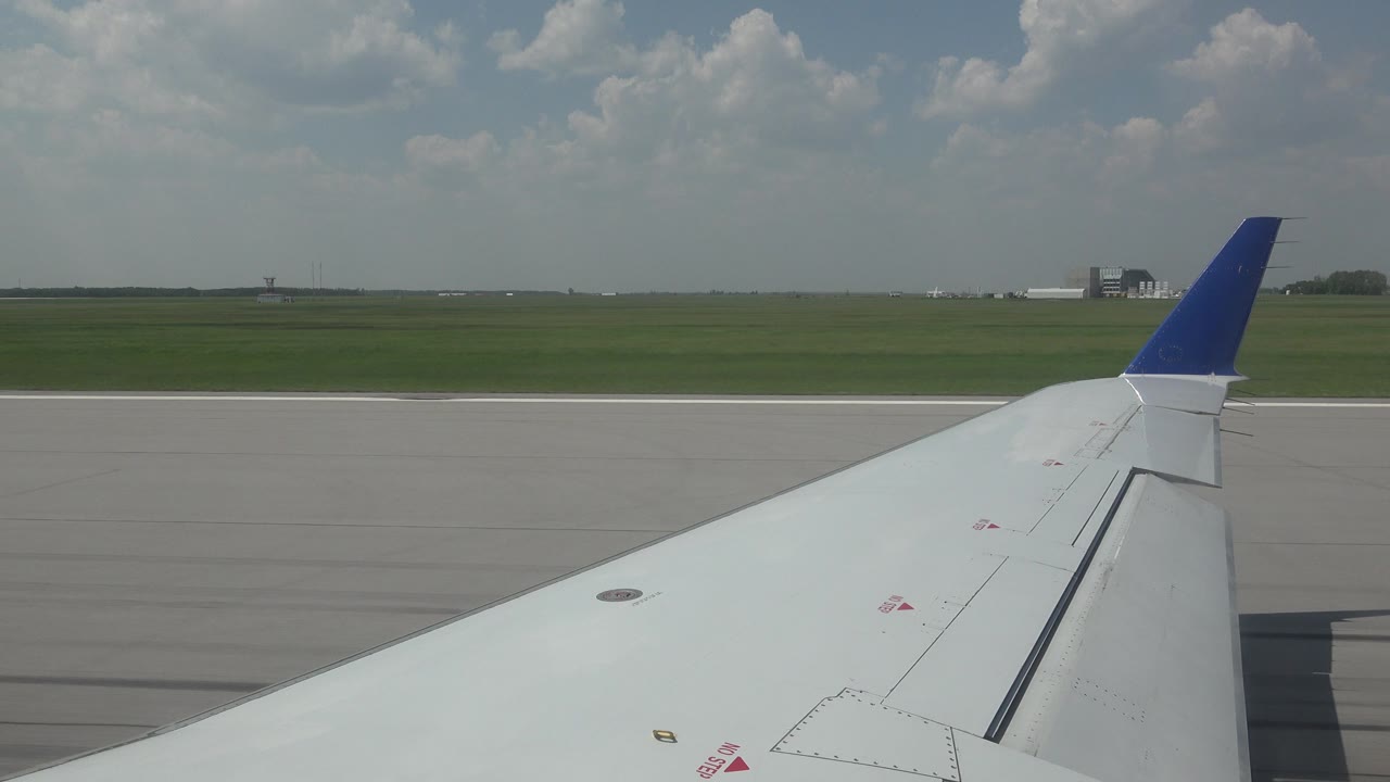 A white airplane wing with a blue winglet is seen from a passenger-window angle on a Winnipeg runway, with green grass, distant airport structures, clouds, and no visible people.