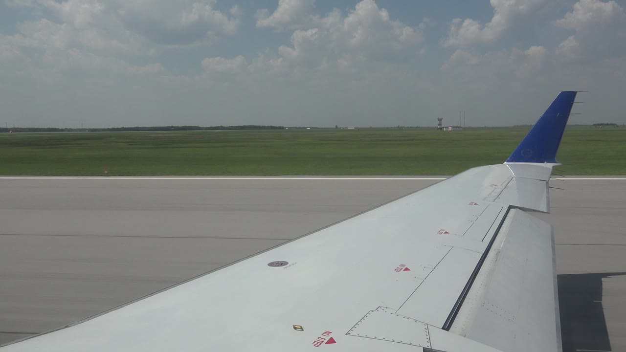 Passenger-window view in Winnipeg showing a white airplane wing with blue tip beside runway pavement, grass, distant horizon, scattered clouds, and no visible people.