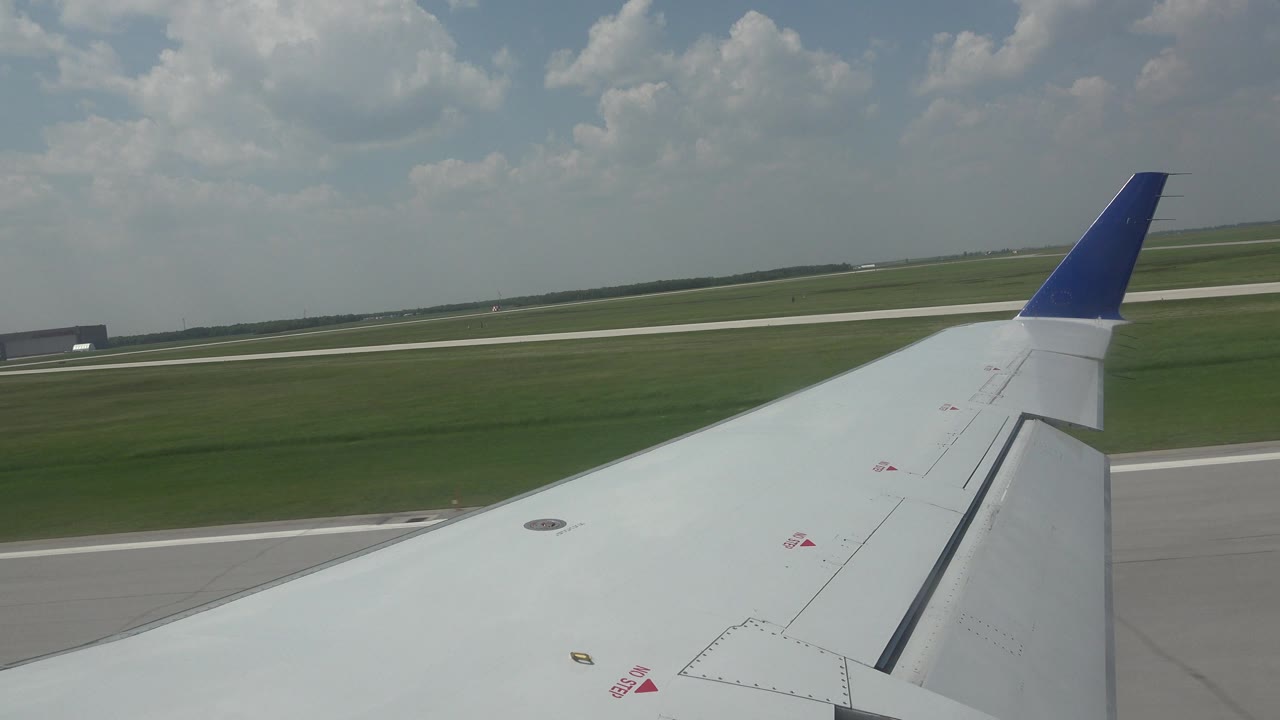 Airplane wing over runway grass under scattered clouds.