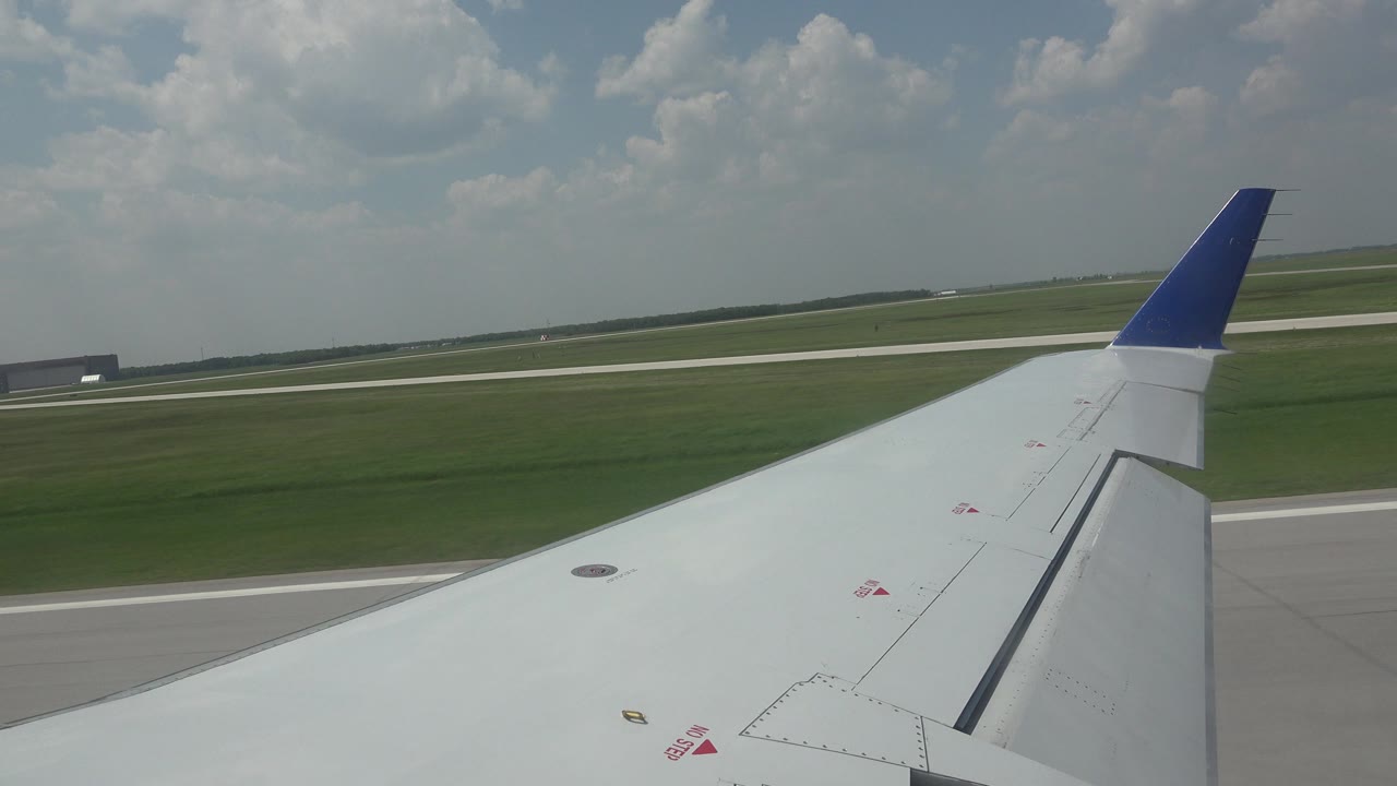 The white wing and blue winglet remain dominant while a runway strip and airport grass sit beyond the window view in Winnipeg, Canada, with no visible people.