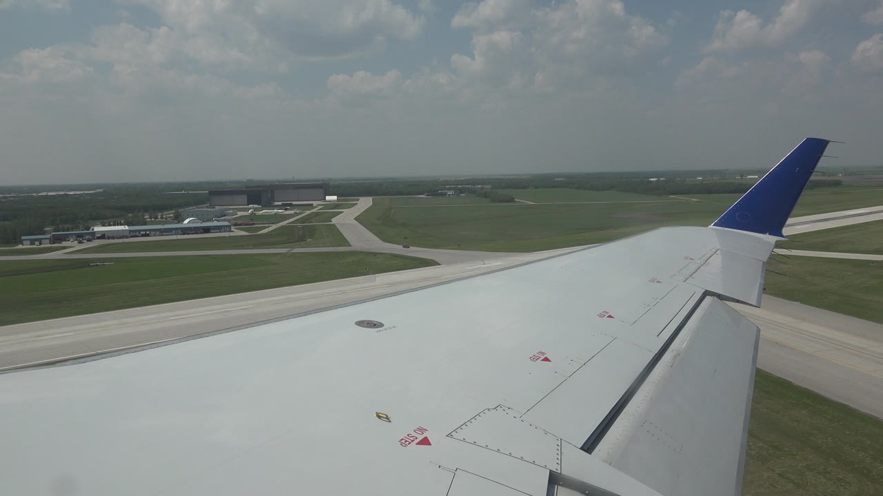 Airplane wing with a blue-tipped winglet over runway pavement and green airport fields in Winnipeg, Canada, with no visible people.