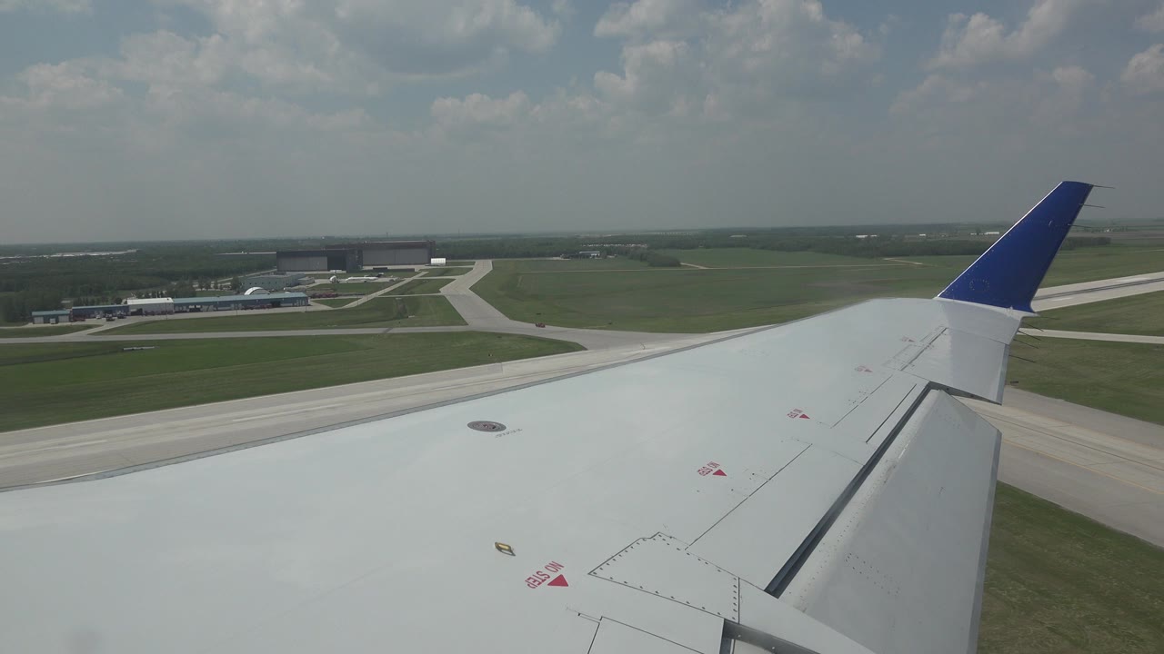 White airplane wing remains in the foreground as airport buildings, paved taxiways, and green fields stretch into a hazy Winnipeg horizon, with no visible people.