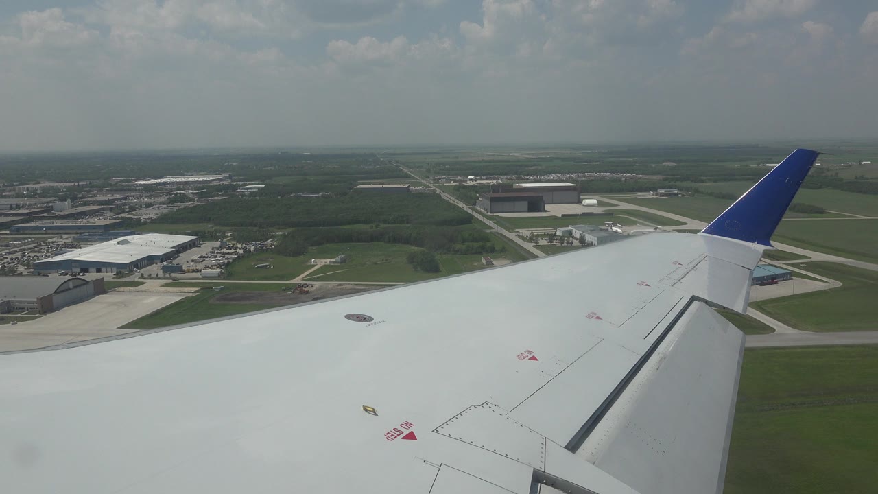 White airplane wing with a blue tip over Winnipeg airport buildings, roads, fields, and hazy sky, with no visible people.