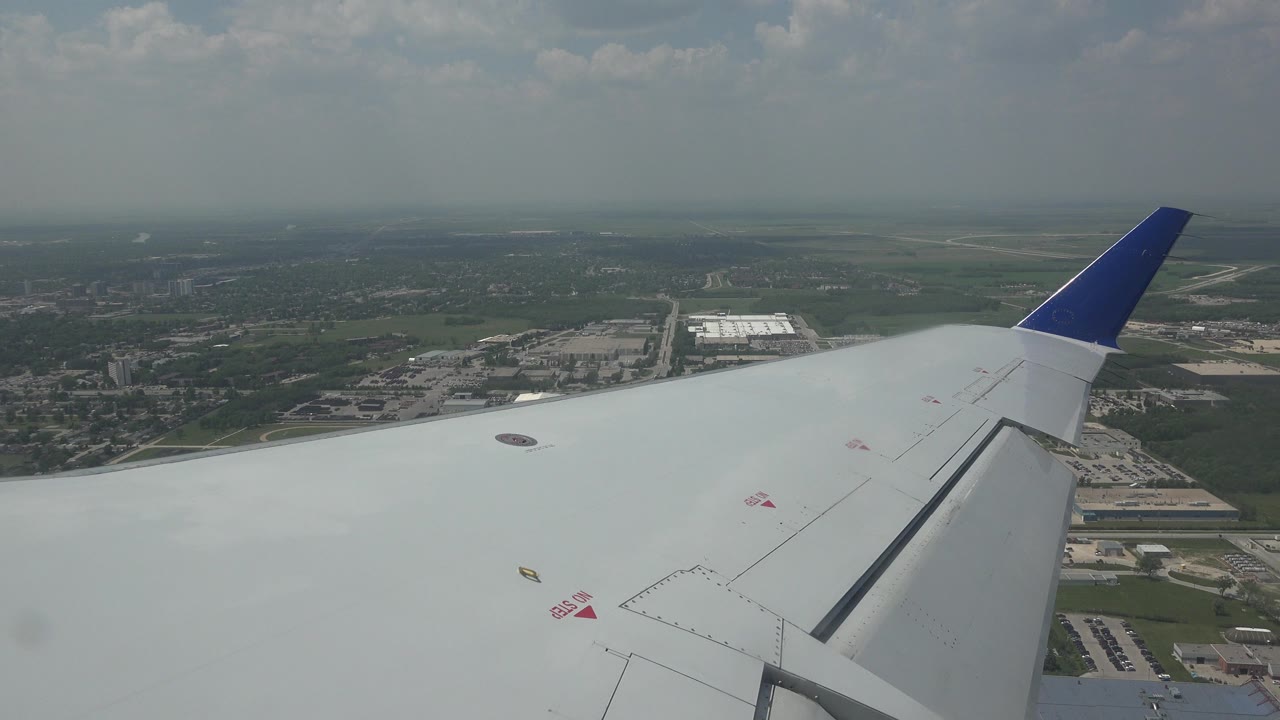 Airplane wing over fields and buildings under a hazy sky.