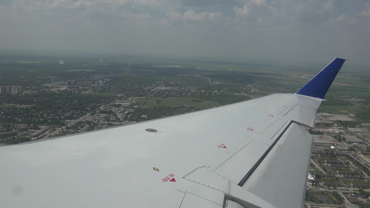 White airplane wing with a blue wingtip over hazy Winnipeg, Canada, with roads, green land, scattered clouds, and no visible people.