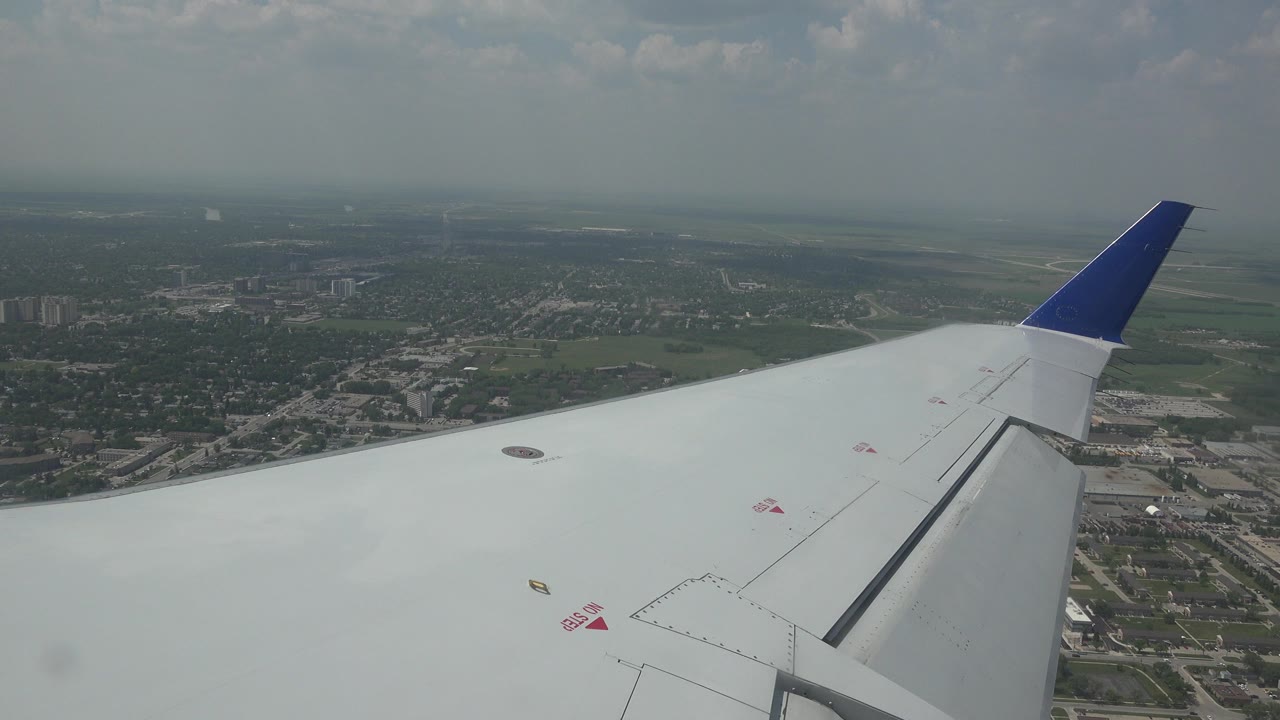 Airplane wing seen from a passenger-window angle above Winnipeg, Canada, with suburban blocks, fields, haze, and no visible people.