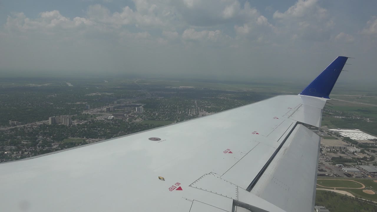 White airplane wing with a blue tip seen over Winnipeg, Canada, with hazy clouds, distant roads, buildings, green land, and no visible people.