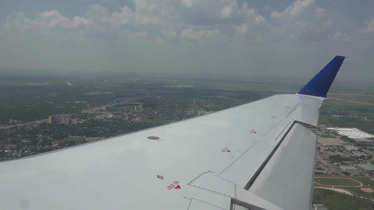 Passenger-window view of a white airplane wing over Winnipeg, Canada, with the wingtip near the upper right, scattered clouds, and no visible people.