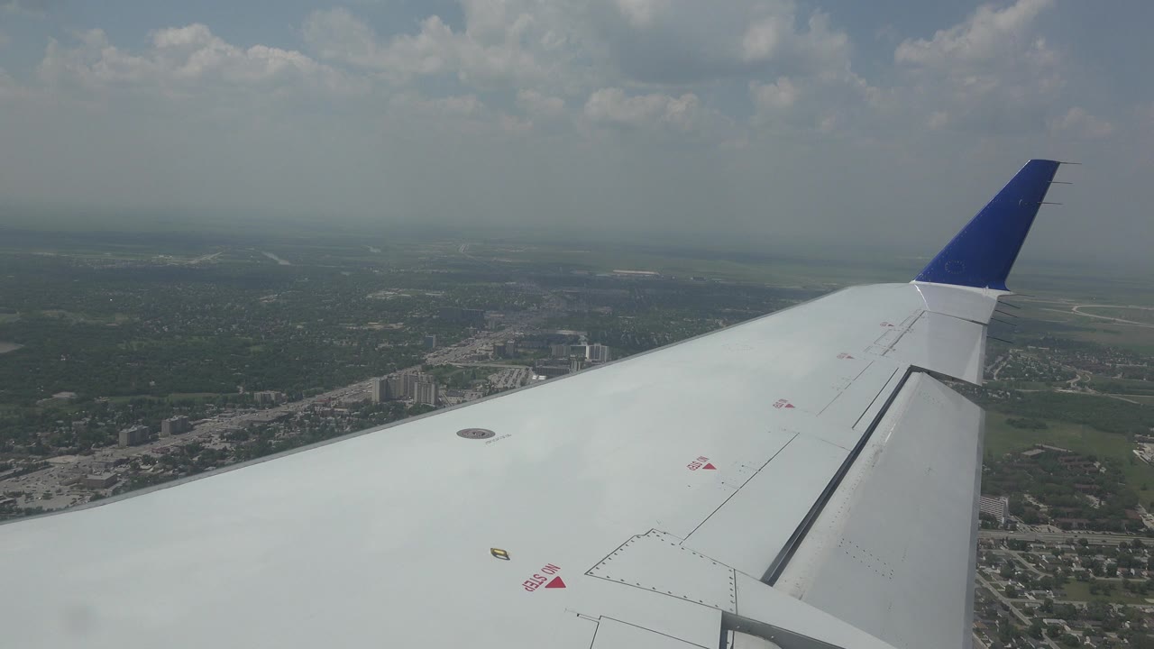 Passenger-window view over Winnipeg, Canada, with a white airplane wing in the foreground, blue wingtip, hazy sky, city blocks, and green land below, with no visible people.