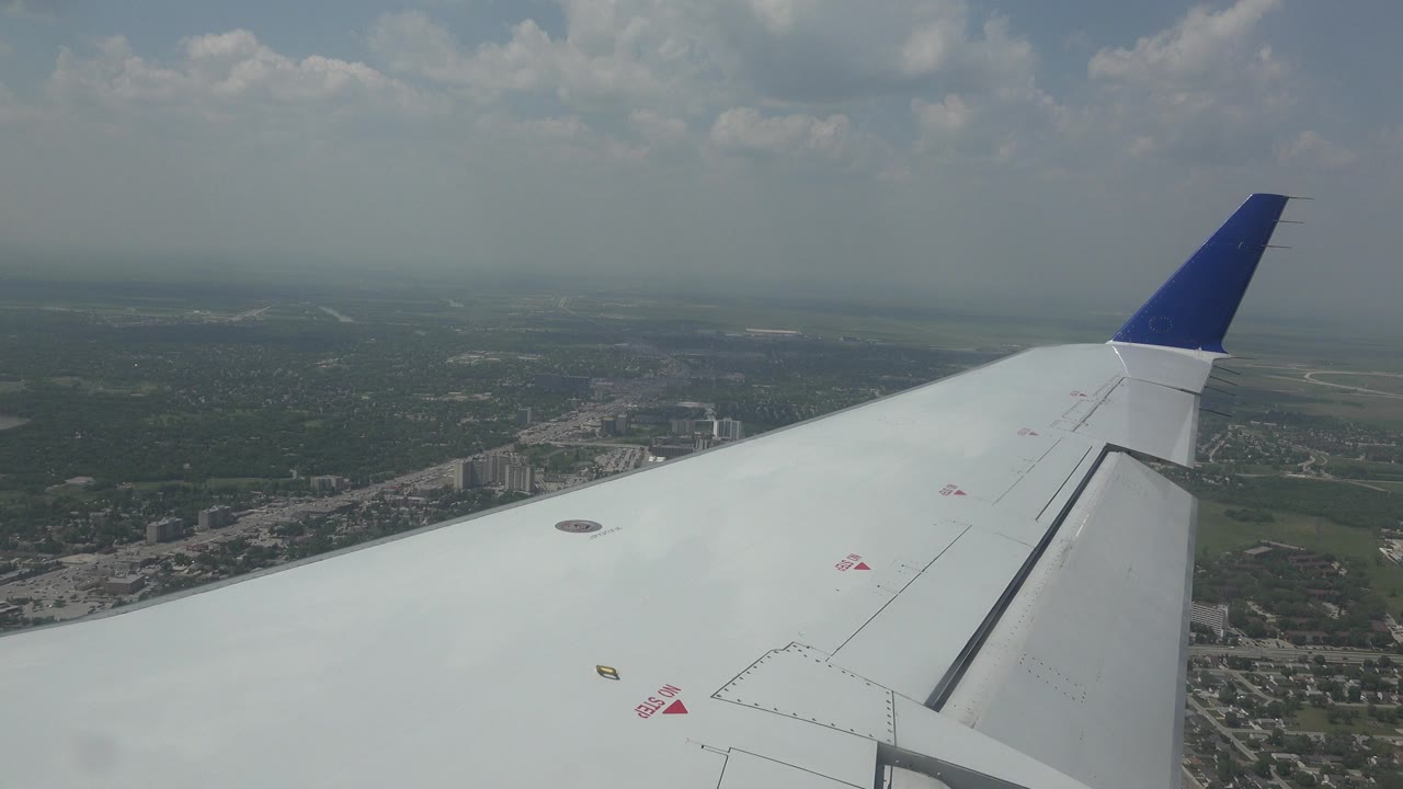 White aircraft wing and blue wingtip seen from inside a plane over Winnipeg, Canada, with roads, buildings, fields, and a hazy cloud line in the distance, with no visible people.