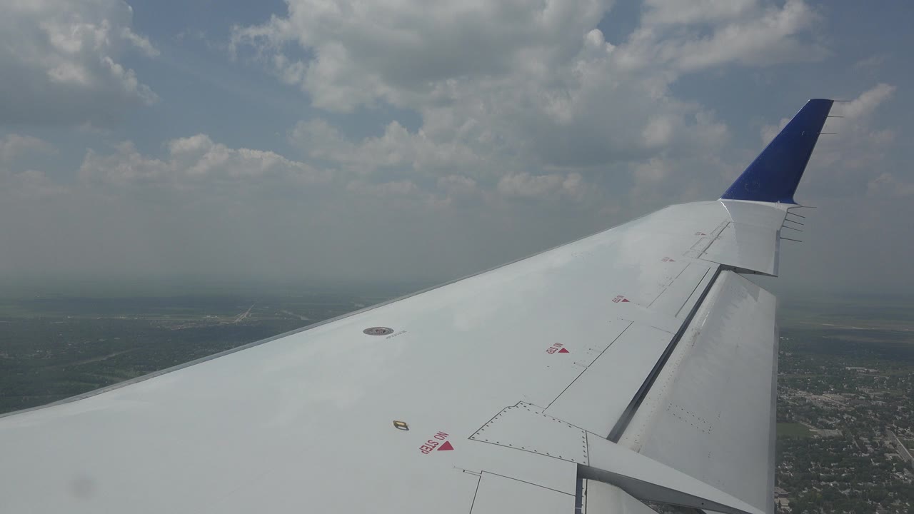 A white aircraft wing stretches across the foreground over Winnipeg, Canada, with a blue wingtip near the upper frame, soft clouds, distant terrain, and no visible people.