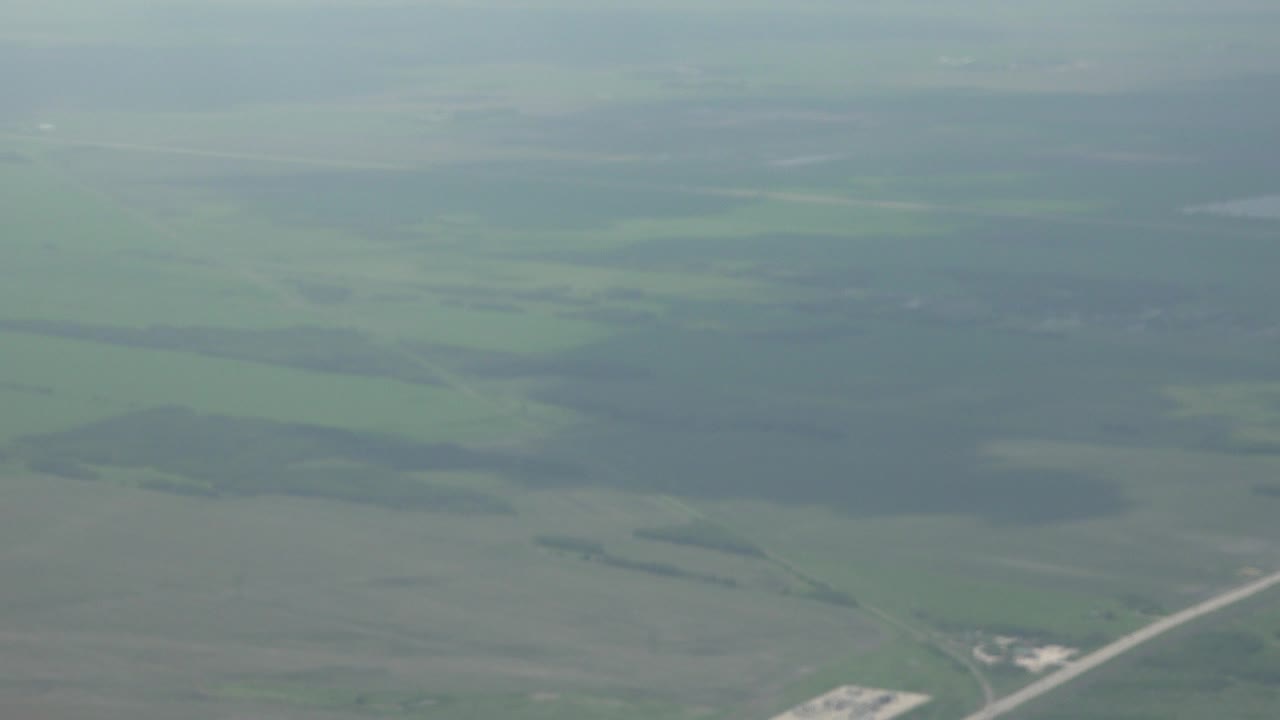 Hazy aerial view near Winnipeg, Canada, looking over green farmland, darker wooded patches, and a pale road near the lower edge, with no visible people.
