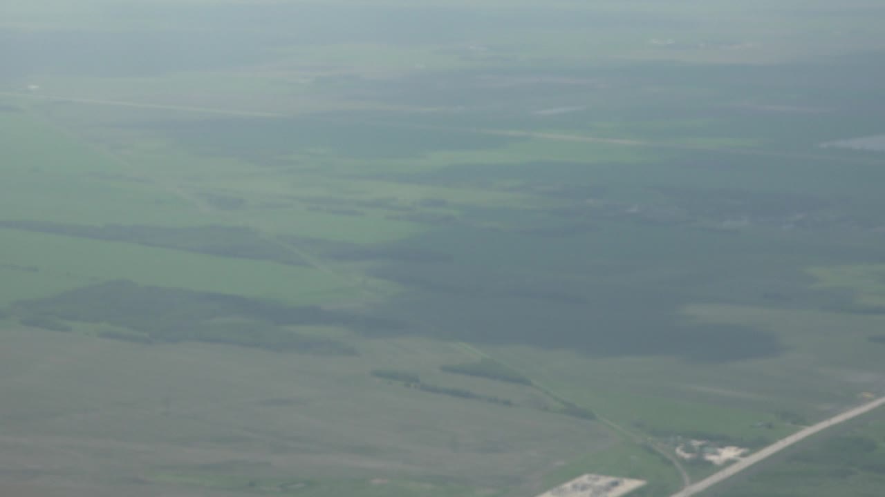 A hazy Winnipeg, Canada aerial frame shows broad green fields, scattered dark tree blocks, and a faint diagonal road, with no visible people.