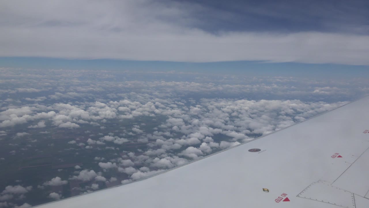 Opening frame of a white airplane wing over scattered cloud tops and dark land below near Winnipeg, Canada, with no visible people.