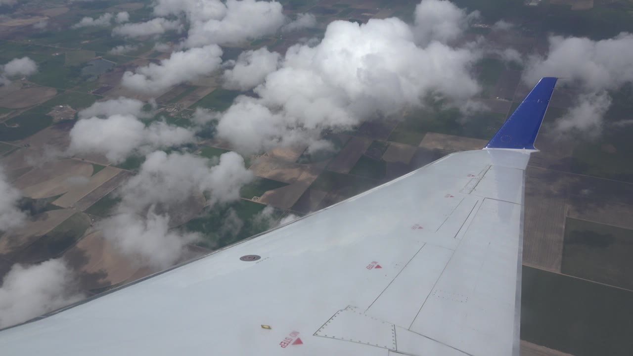 White airplane wing with a blue tip seen from a window seat above clouds and farmland near Winnipeg, Canada, with no visible people.