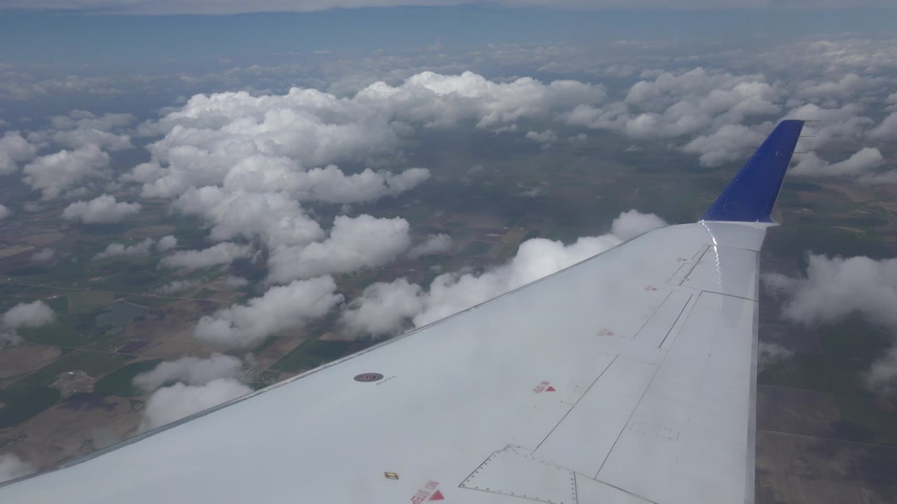 A white airplane wing with a blue wingtip extends over scattered clouds and patchwork land near Winnipeg, Canada, with no visible people.