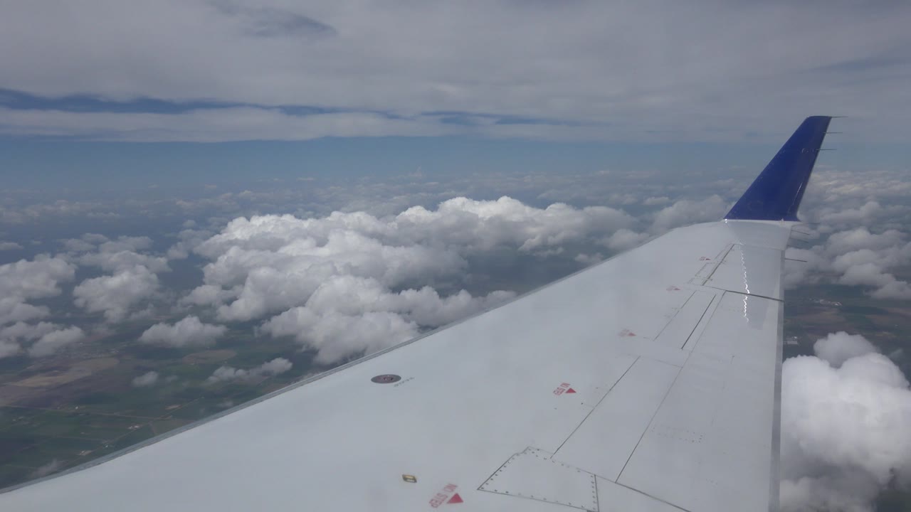 White airplane wing with a blue wingtip seen from a passenger window over Winnipeg, Canada, with clustered clouds and faint terrain below, no visible people.