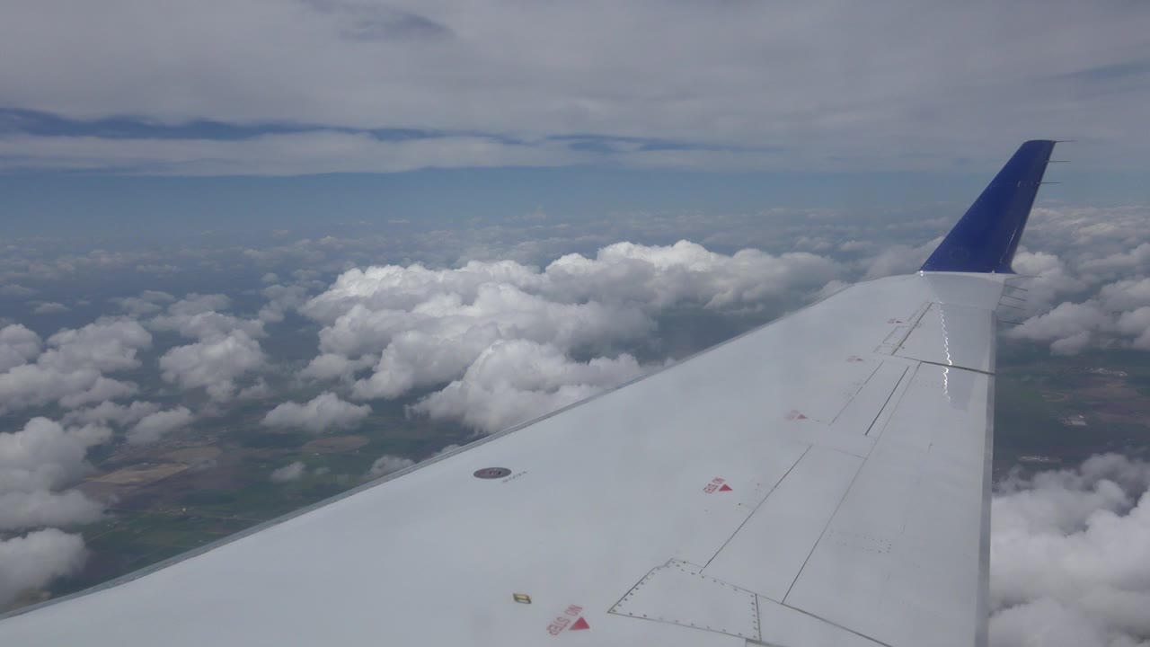 Opening view from an airplane window over Winnipeg, Canada, with a white wing and blue winglet above clouds and distant ground, no visible people.