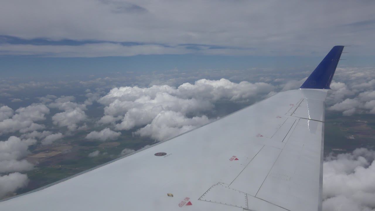 A white aircraft wing remains prominent over Winnipeg, Canada, with bright cloud clusters and faint ground patterns visible beyond it, no visible people.