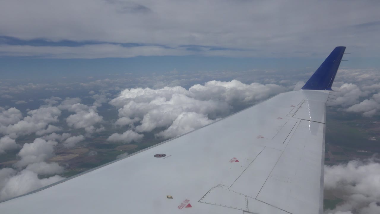 A white airplane wing with a blue tip extends over scattered clouds near Winnipeg, Canada, with no visible people.