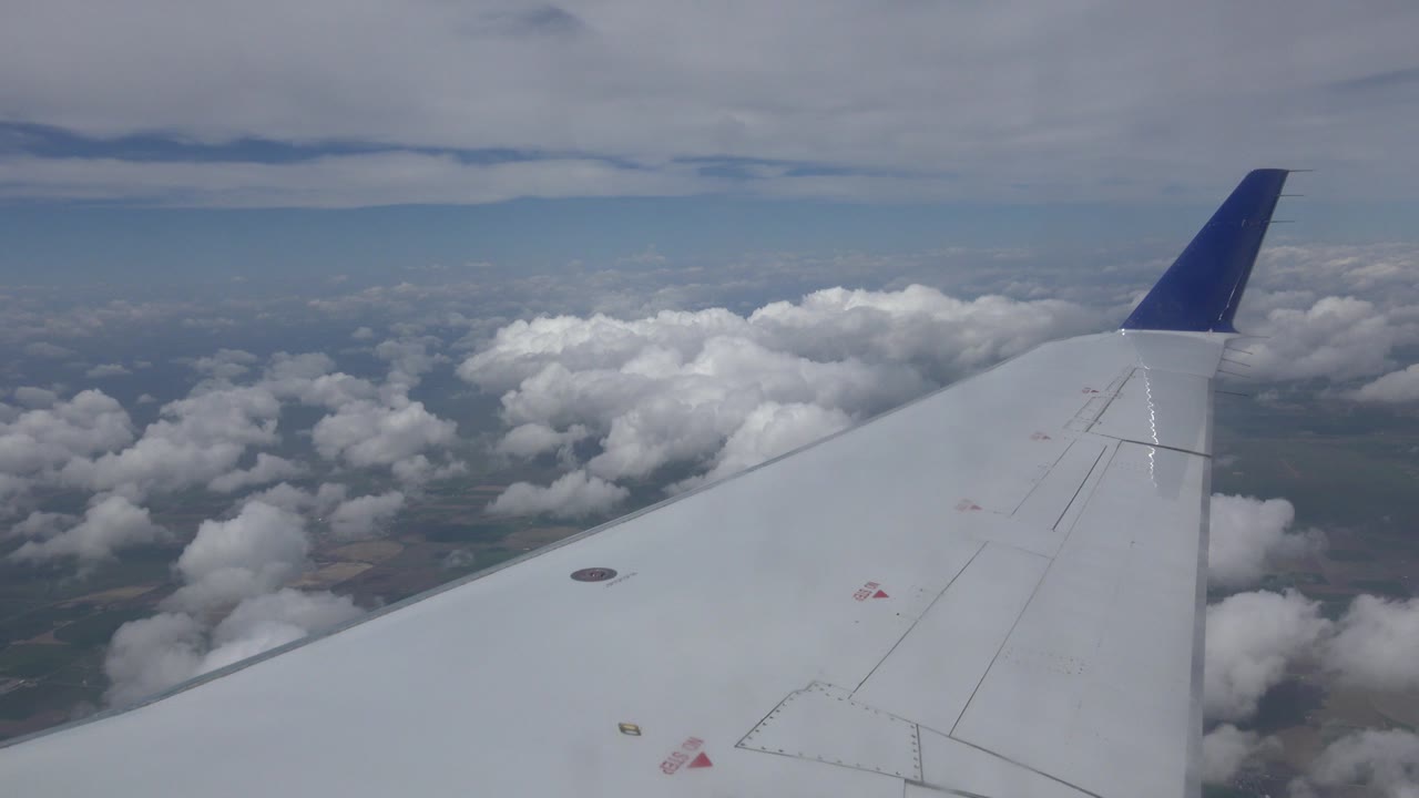 Passenger-window view near Winnipeg, Canada, with a pale airplane wing and blue winglet above scattered clouds, no visible people.