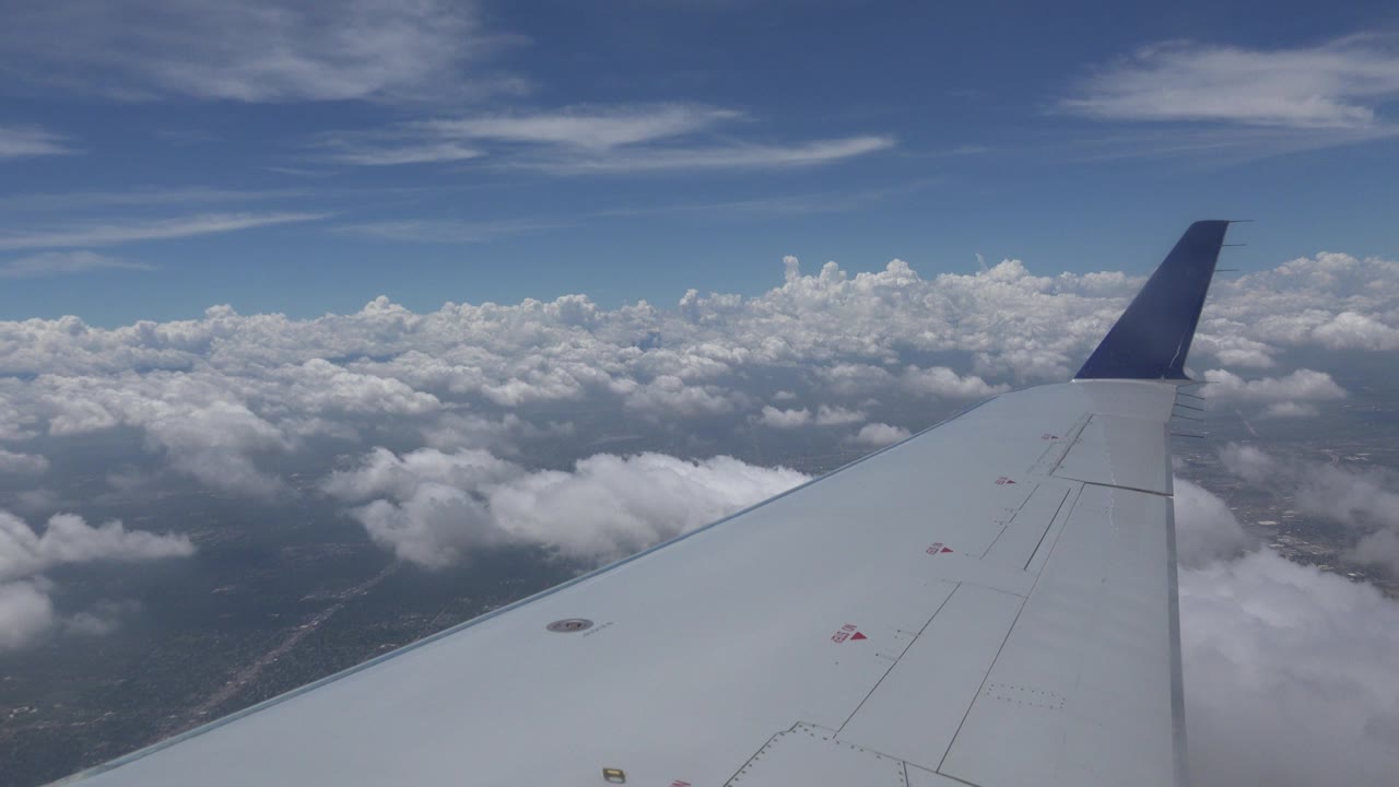 Passenger-window view near Winnipeg, Canada with a white airplane wing and dark blue wingtip over scattered clouds, blue sky, and distant land, with no visible people.
