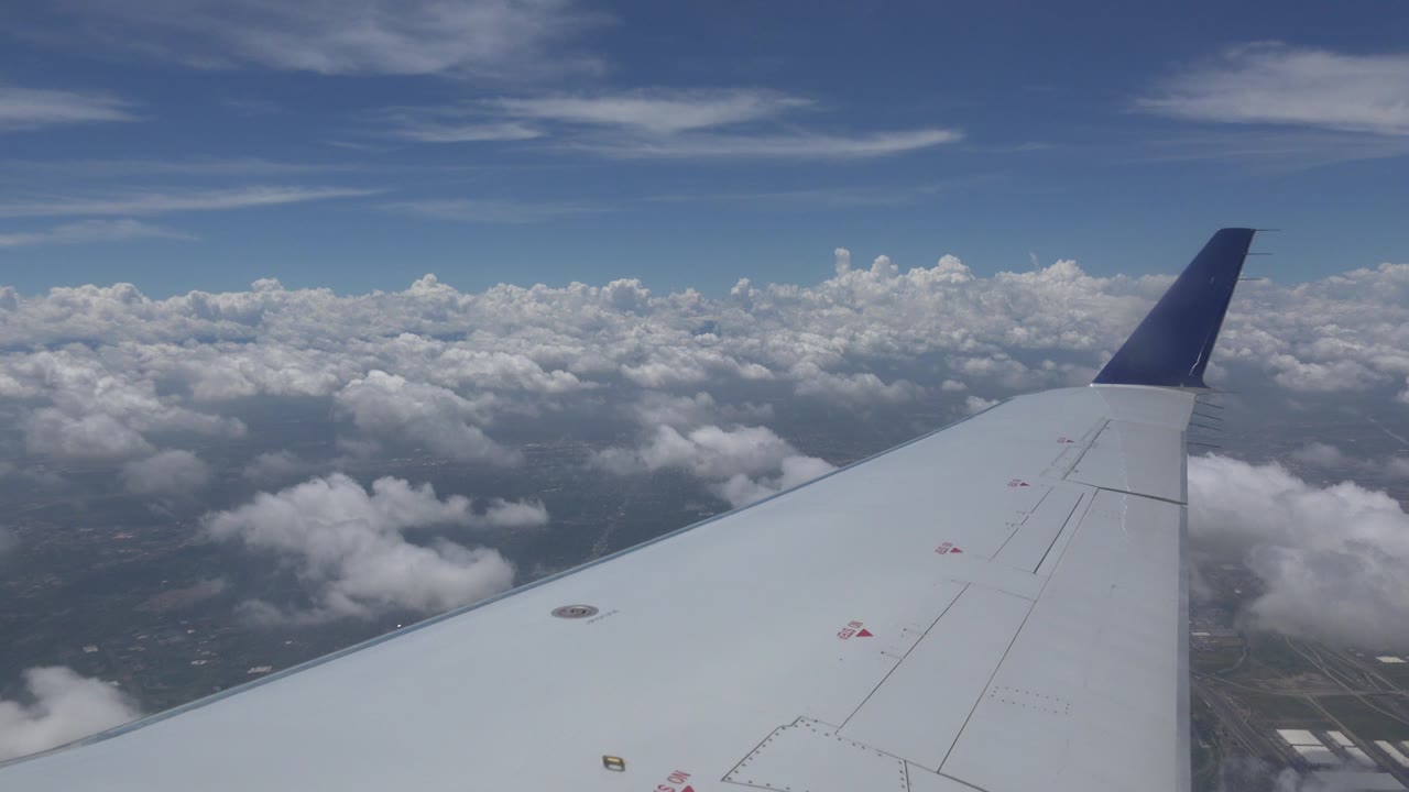 At 0:00, a white airplane wing with a dark blue tip stretches across a Winnipeg, Canada passenger-window view above scattered clouds and faint ground below, with no visible people.