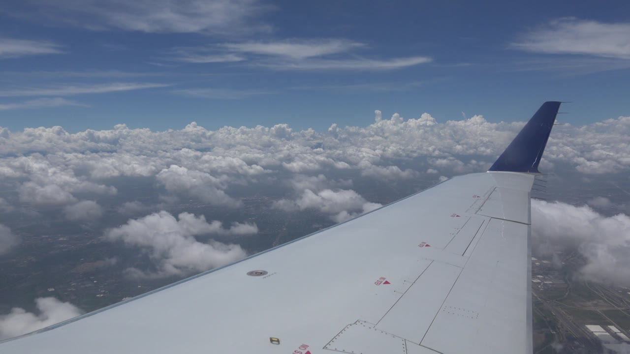 Airplane wing and blue wingtip above clouds and distant land.