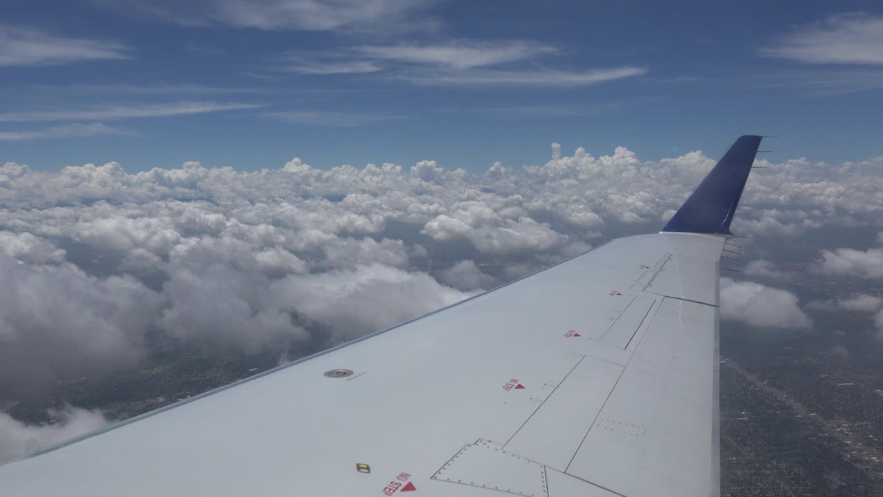 At 0:01, the plane wing remains centered over dense white clouds with faint ground texture below near Winnipeg, Canada, and no visible people.