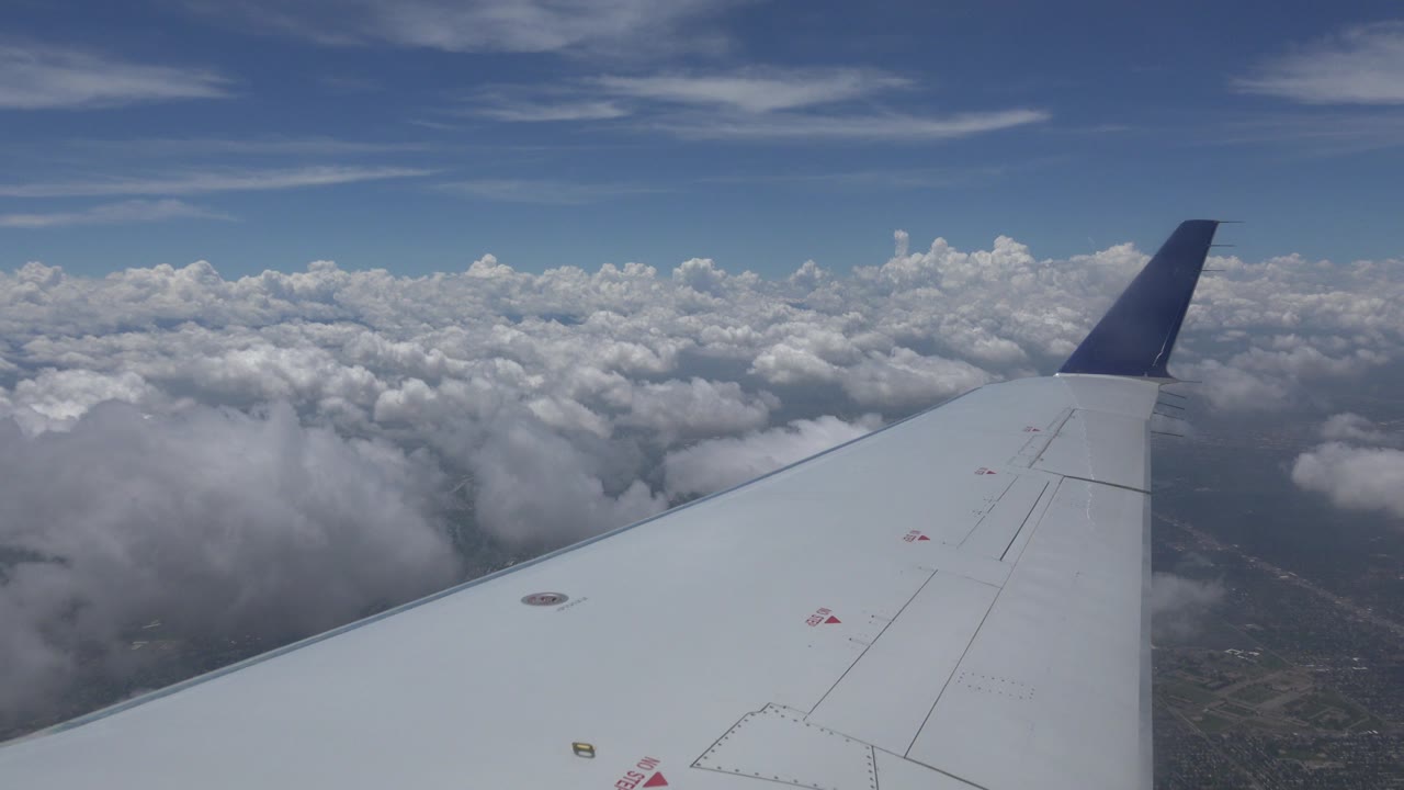 Passenger-window view over Winnipeg, Canada, with a white airplane wing angled toward a dark blue winglet, broken clouds below, blue sky above, and no visible people.
