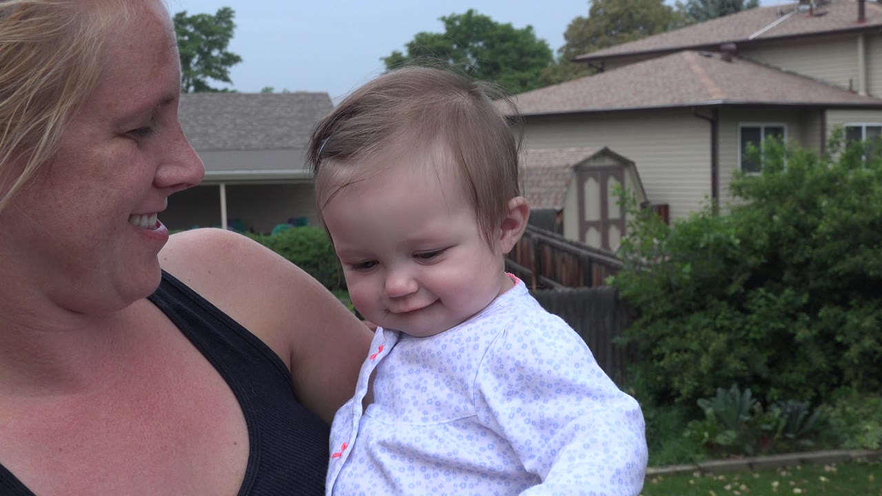 A blonde woman holds a baby girl close in Westminster, Colorado, with shrubs, fencing, and private homes visible behind them.