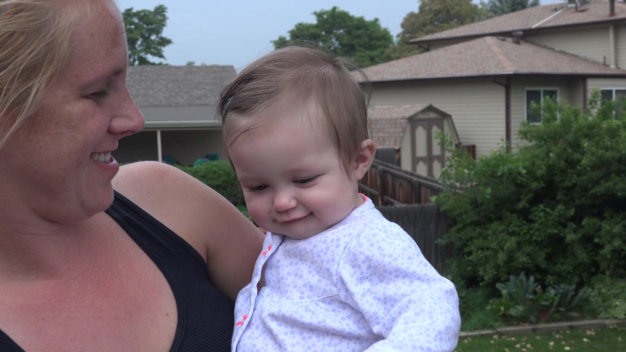 The woman smiles down toward the baby girl in Westminster, Colorado, while the child looks downward against a backdrop of homes and greenery.