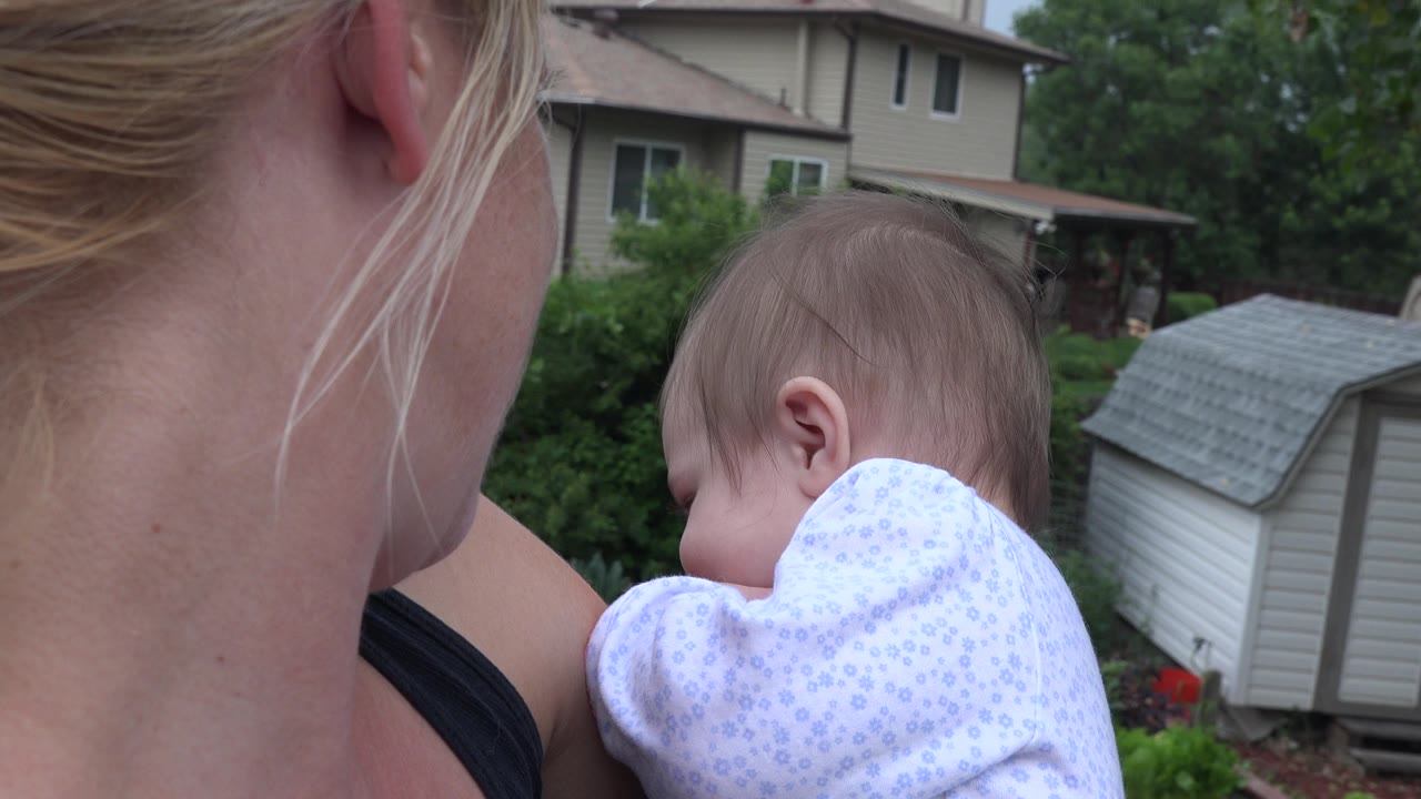The baby turns slightly downward in the woman's arms while gray storage sheds, garden greenery, and a nearby house remain visible in the Westminster backyard.