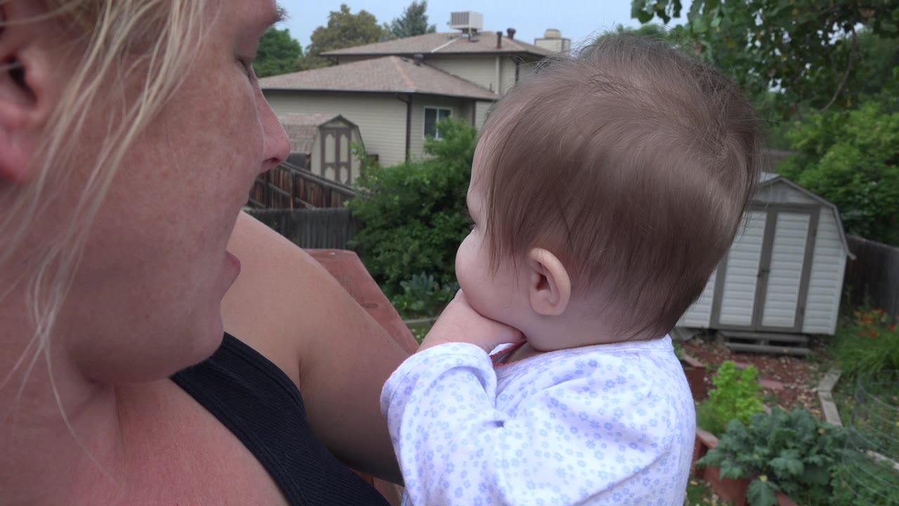 In Westminster, Colorado, a blonde mother holds a baby close beside a backyard deck, with garden plants, a fence, sheds, trees, and houses behind them.