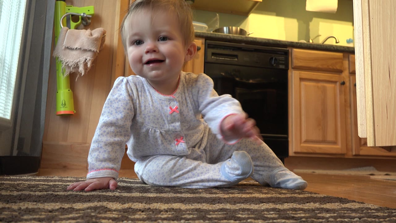 Toddler girl remains seated on the rug and reaches one hand forward toward the floor-level camera inside the Westminster kitchen.