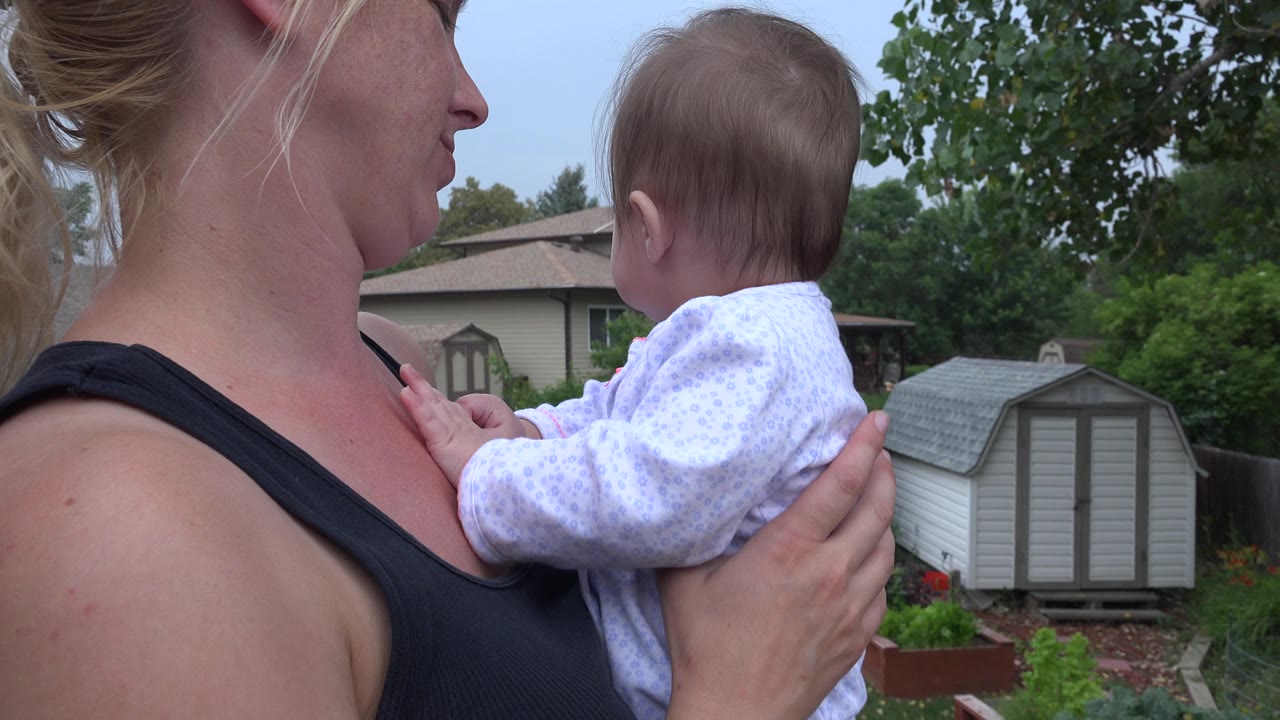 The woman continues holding the baby close to her chest while the baby looks toward the yard, with a gray shed, fence, trees, and houses in the background.