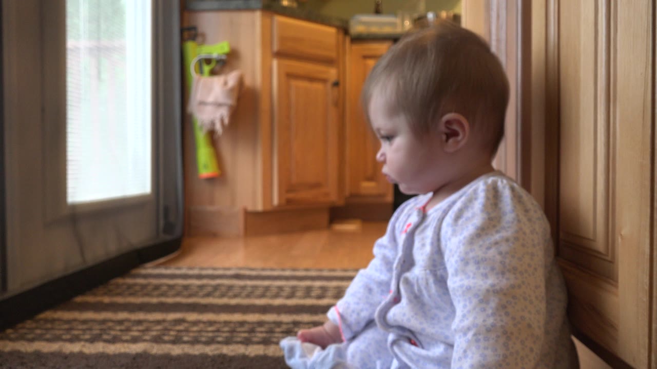 Baby seated beside wood trim on a striped rug near kitchen cabinets.