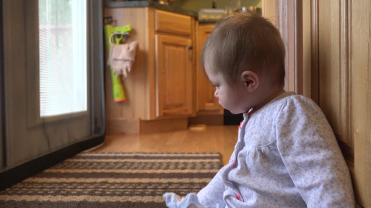 At 0:02, the baby shifts farther right against the wood trim, with the striped rug leading back toward the honey-colored kitchen cabinets.