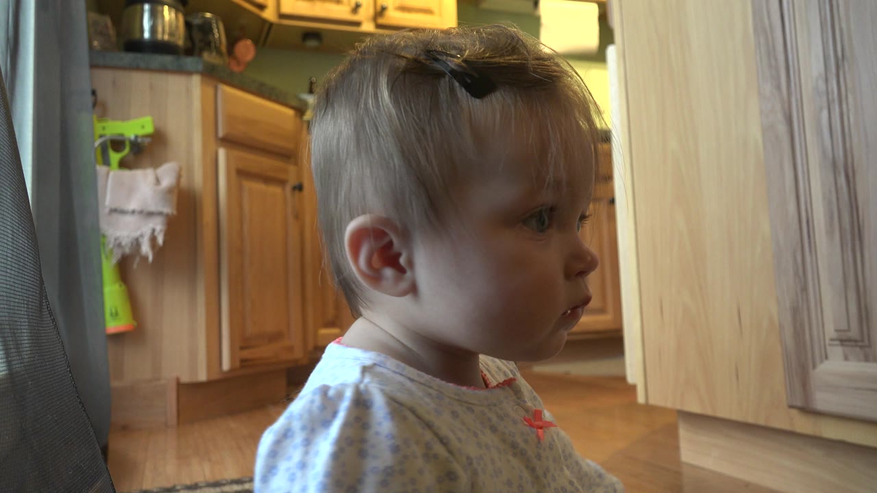 The baby girl remains in side profile in the Westminster kitchen, with her face turned right and the cabinet line visible across the background.