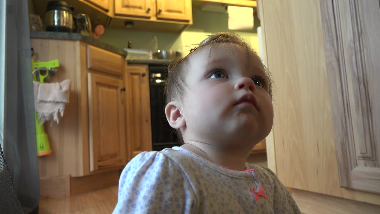 The baby continues looking upward near the camera, with a pale vertical doorway or cabinet edge framing the right side of the Westminster kitchen scene.