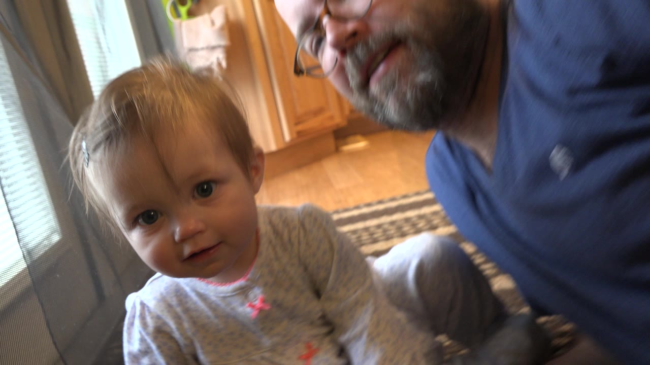 The Westminster home interior shows the infant centered closer to the camera, with the adult man's partial face and torso at the right edge and bright door glass behind them.