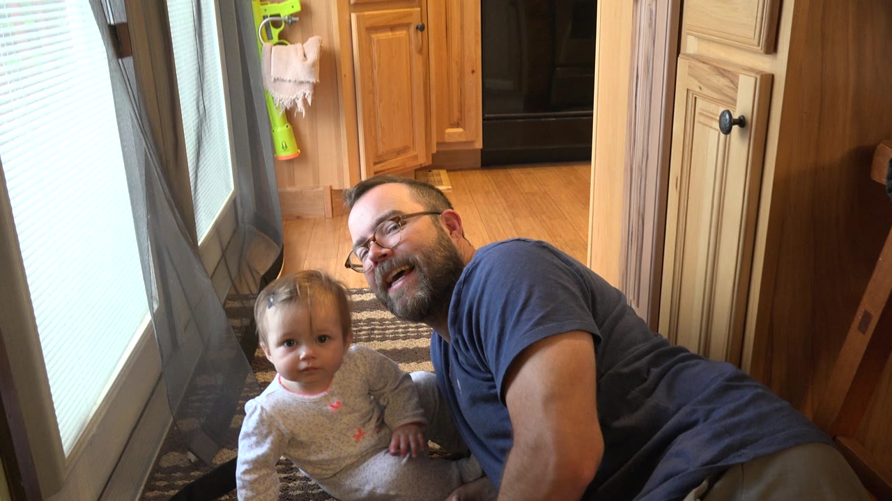 The toddler looks forward from the rug while the smiling father stays low beside her, with warm wood cabinets and the door area behind them in Westminster, Colorado.