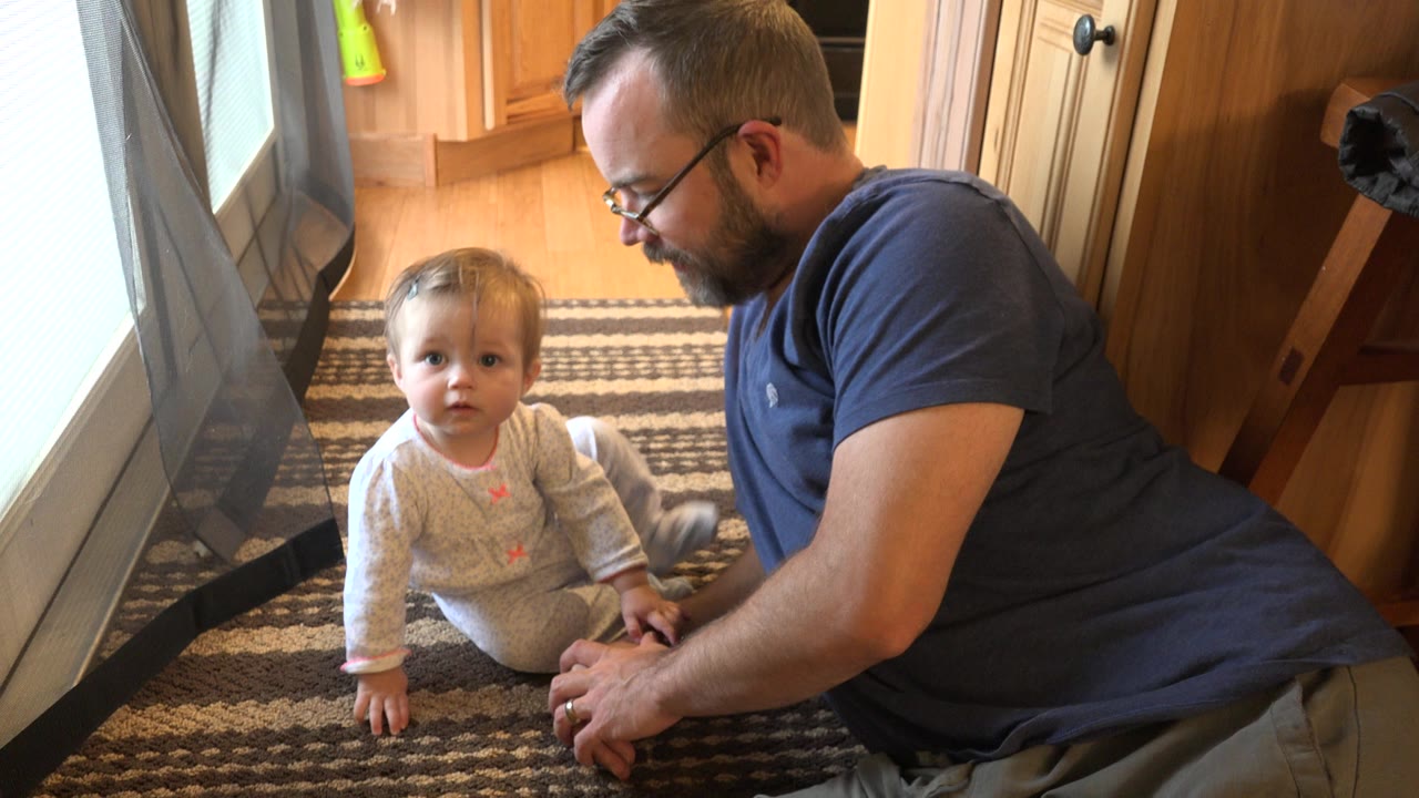 Father lying on a rug beside a seated toddler near a bright window.