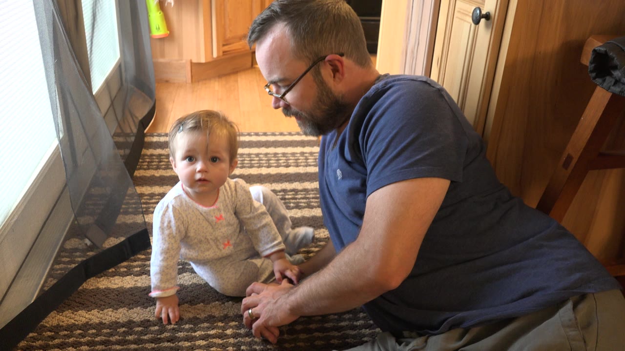 In Westminster, Colorado, the toddler looks toward the camera while the father stays close on the rug near the window and curtain.